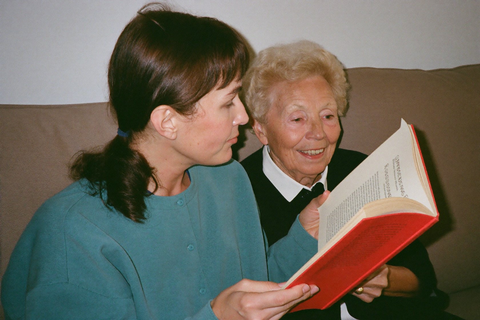 A caregiver reading a book beside a senior woman on the couch
