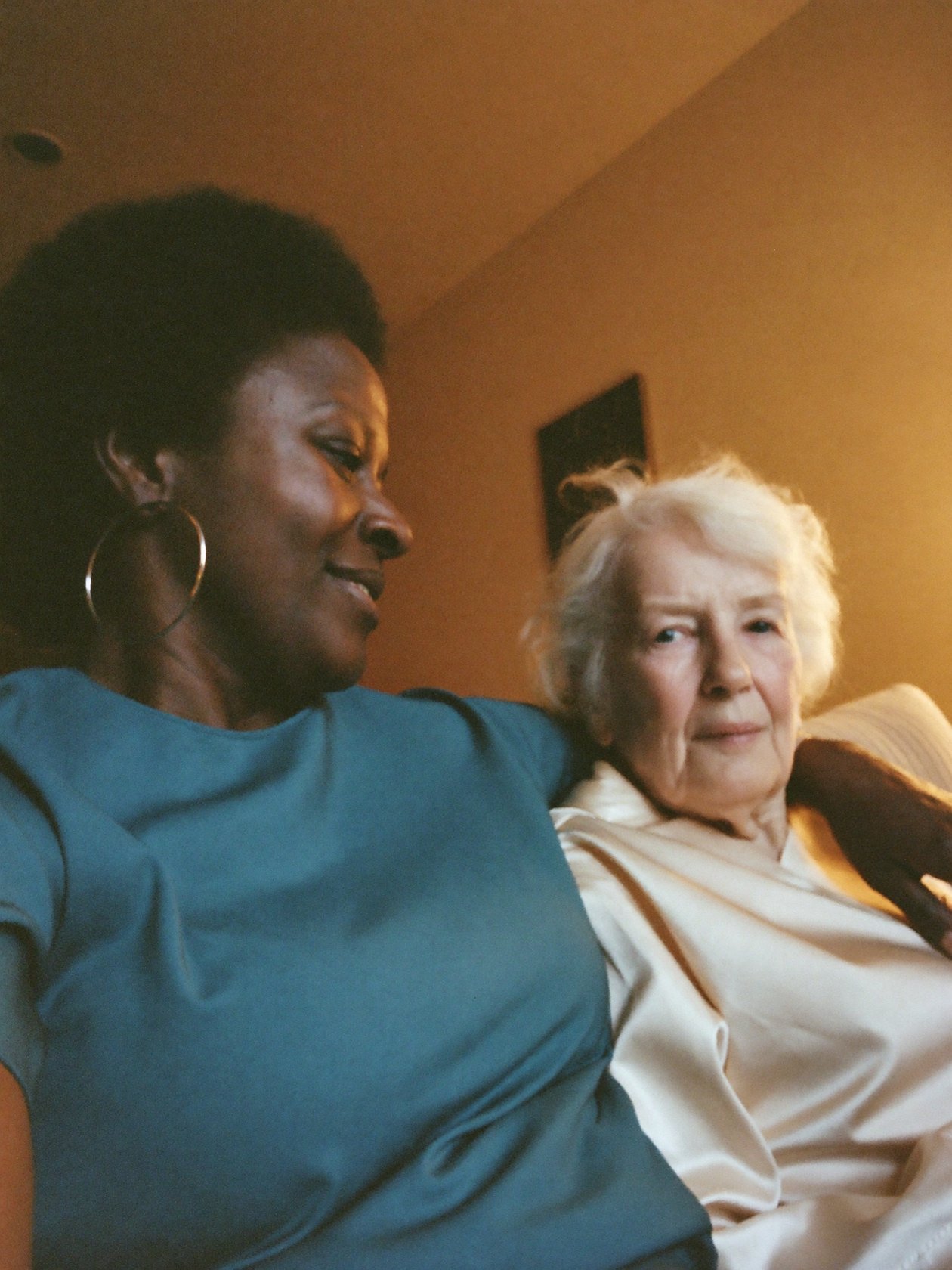 A caregiver sitting with a senior woman in a lamplit living room in the evening