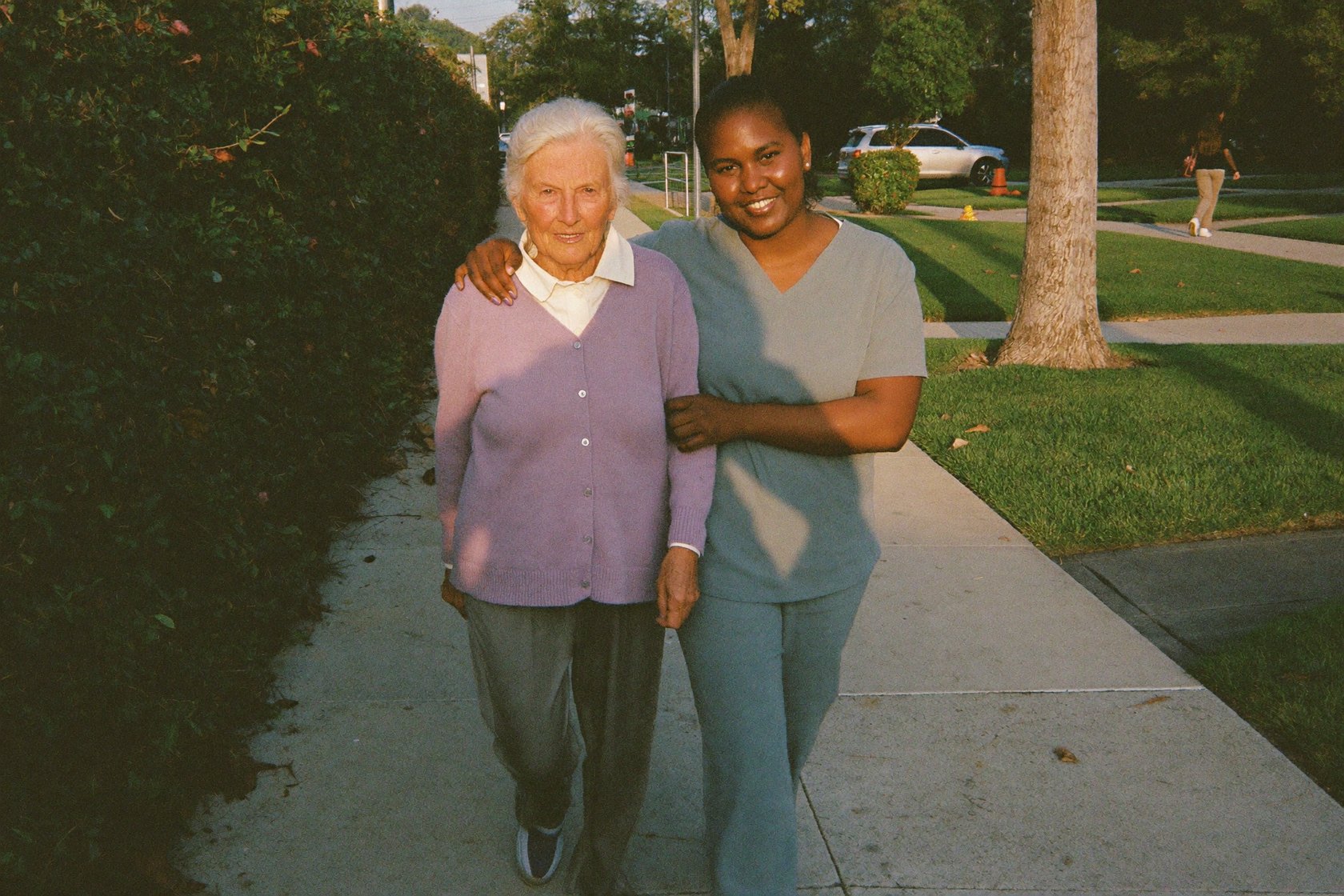 A senior woman walking arm-in-arm with her caregiver along a tree-lined sidewalk