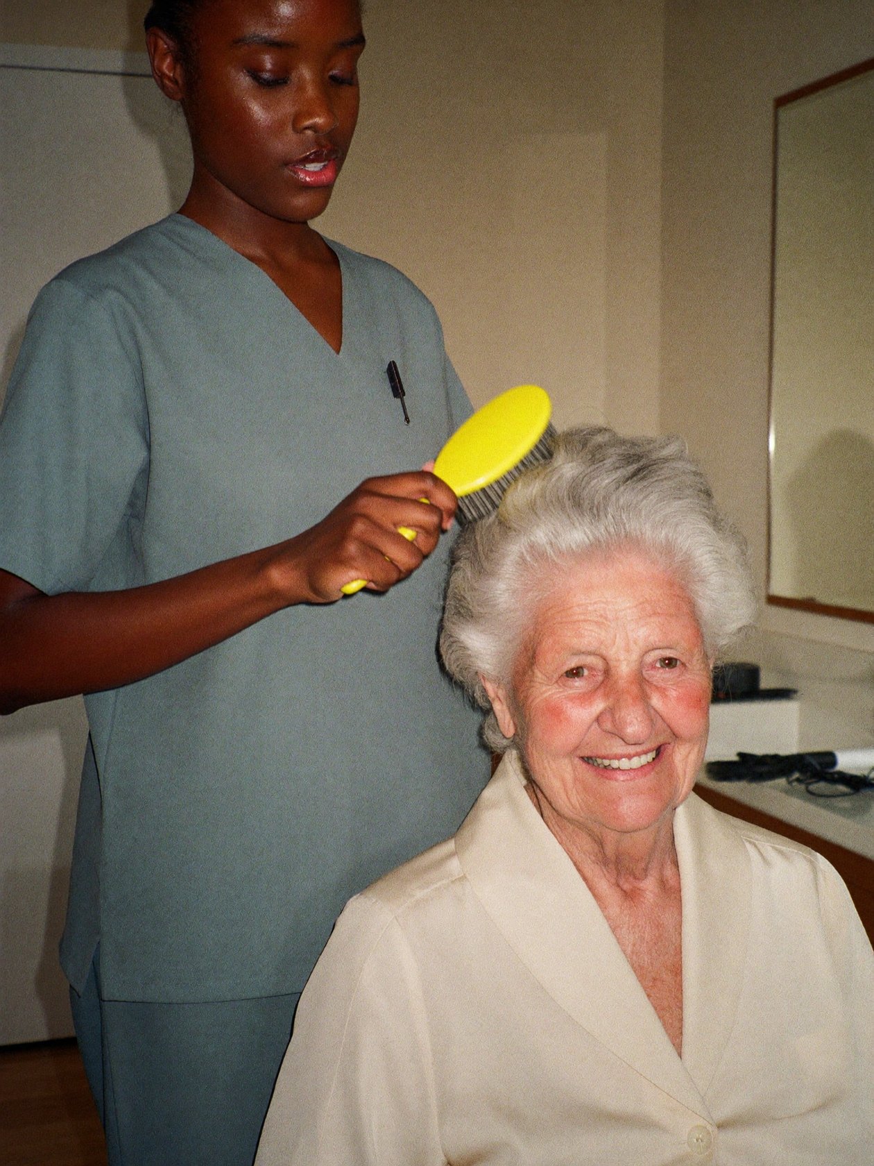 A caregiver brushing and styling a senior woman's hair at her vanity