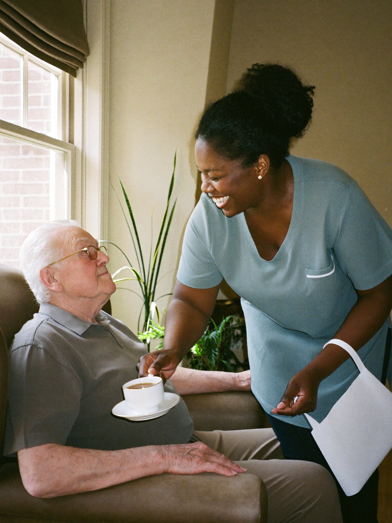 A caregiver bringing a cup of tea to a senior man in his armchair